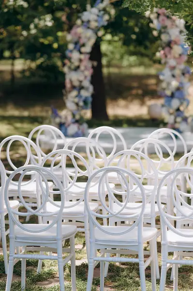 Sırtında yuvarlak sırtı olan beyaz modern sandalyeler açık havada uzun ağaçların arasında kumlu bir zemin üzerinde duruyor.