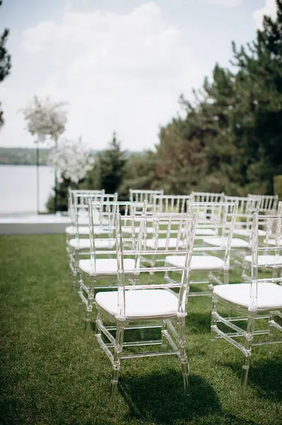 Beyaz yastıklı sıra sıra sandalyeler açık havada bir düğün ortamında göle bakan yeşil bir çimenlikte duruyor.