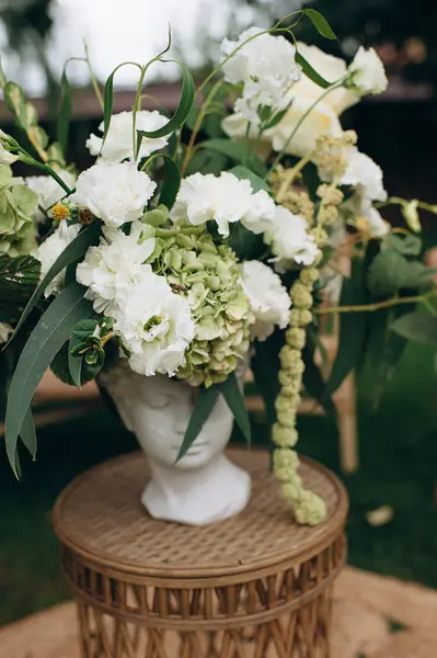 Bir vazonun içindeki beyaz bir çiçek aranjmanı, bahçedeki mobilyaların arasında dokuma bir masanın üzerinde duruyor.