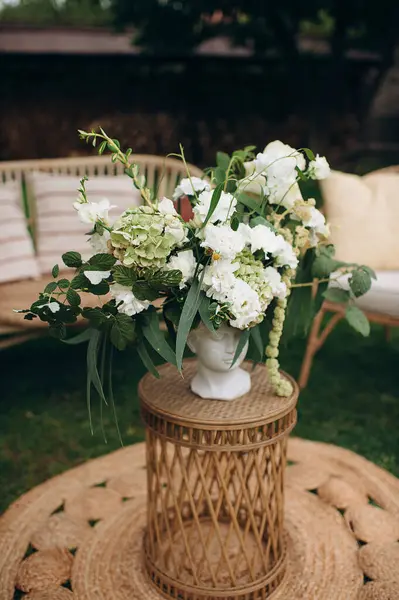 Bir vazonun içindeki beyaz bir çiçek aranjmanı, bahçedeki mobilyaların arasında dokuma bir masanın üzerinde duruyor.