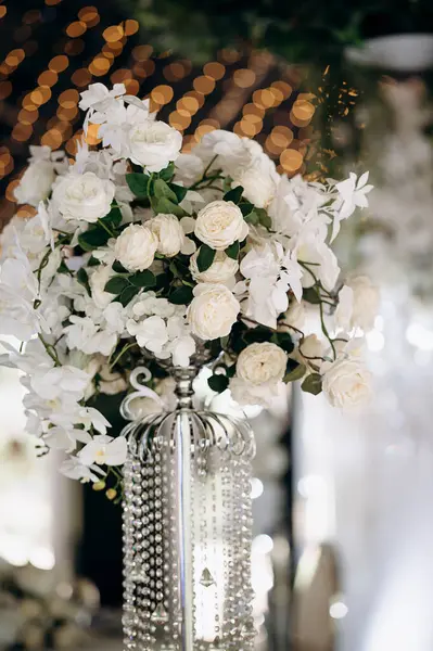 Beyaz gülleri ve orkideleri olan uzun bir çiçek süsü bir etkinliğin ortasında aynalı bir masada duruyor.