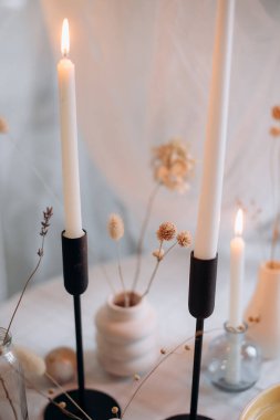 Arka planda kurumuş çiçekleri olan siyah tutacaklı iki beyaz mum yakın plan.