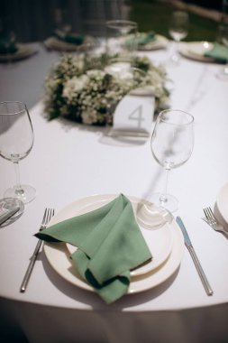 Yeşil peçeteli, beyaz tabaklı, zarif yuvarlak masa takımı ve masa numarası olan bir süs eşyası.