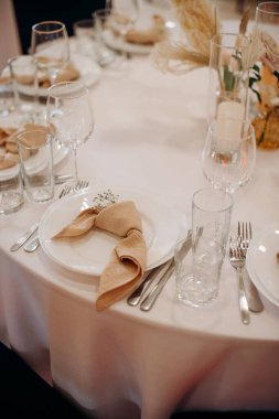 Bej düğümlü bir peçete ve beyaz masa örtüsü üzerinde narin bir çiçek aksanıyla zarif yuvarlak masa seti.