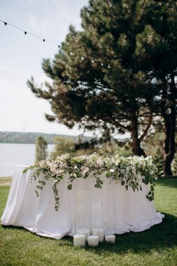 Beyaz masa örtüsü, okaliptüs çelengi ve sütun mumlarıyla şık bir açık hava düğün başkanlığı.