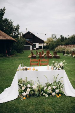 Beyaz masa örtüsü, beyaz çiçek aranjmanları ve şeffaf Chiavari sandalyeleri olan zarif bir açık hava düğün başkanı.