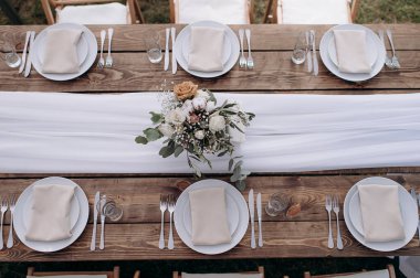 Üzerinde ahşap bir masa, beyaz bir koşucu, beyaz plaka ve çiçek desenli bir düğün masası manzarası var.