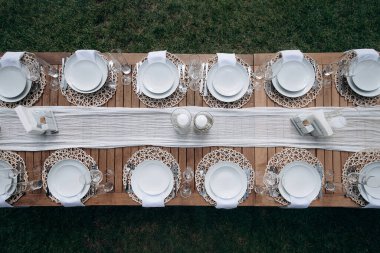 Beyaz tabaklar, peçeteler ve yeşil çimlerin üzerinde beyaz bir koşucuyla zarif bir şekilde kurulmuş ahşap bir düğün masası manzarası.