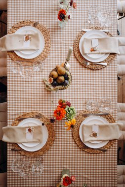 Kırsal bir düğün masası manzarası, kareli bir masa örtüsü, dokunmuş kaplar, beyaz tabaklar ve meyve dekoru.