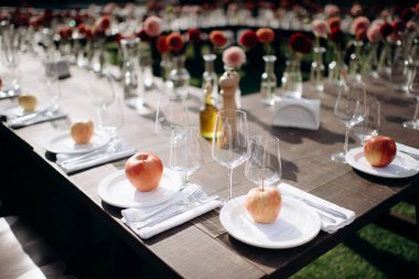 Koyu ahşap bir masa, beyaz tabaklar, iyilik olarak elmalar ve çiçek süslemeleri olan şık bir açık hava düğün masası.