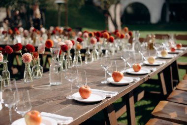 Koyu ahşap bir masa, beyaz tabaklar, iyilik olarak elmalar ve çiçek süslemeleri olan şık bir açık hava düğün masası.