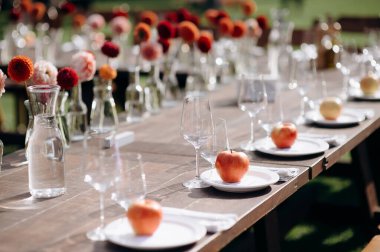 Koyu ahşap bir masa, beyaz tabaklar, iyilik olarak elmalar ve çiçek süslemeleri olan şık bir açık hava düğün masası.