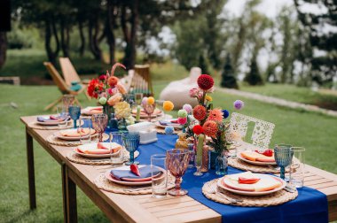 Mavi bir koşucu, dokuma tabaklar, pembe tabaklar ve renkli çiçek süslemeleri olan şık bir açık hava düğün masası.
