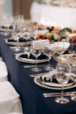 Koyu mavi masa örtüsü, gümüş boncuklu şarj aletleri, beyaz tabaklar ve koyu peçeteli şık bir düğün masası.