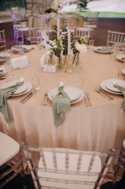 Açık bej masa örtüsü, beyaz tabaklar, adaçayı yeşili peçeteler ve basit çiçek süslemeleri olan zarif bir düğün masası.