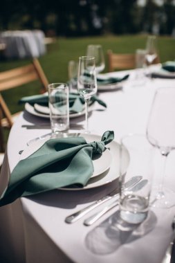 Beyaz masa örtüsü ve beyaz tabakta zümrüt yeşili peçetelerle süslenmiş zarif bir açık hava düğün masasının yakın çekimi.