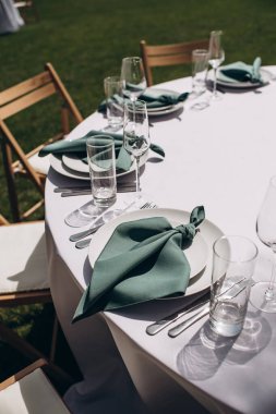 Beyaz masa örtüsü ve beyaz tabakta zümrüt yeşili peçetelerle süslenmiş zarif bir açık hava düğün masasının yakın çekimi.