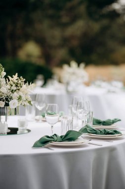Beyaz masa örtüsü ve beyaz tabakta zümrüt yeşili peçetelerle süslenmiş zarif bir açık hava düğün masasının yakın çekimi.