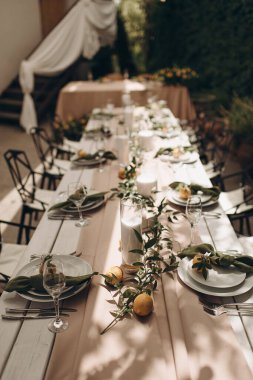 Beyaz ahşap bir masa, açık pembe bir koşucu, beyaz tabaklar ve narenciye aksanlı şık bir açık hava düğün masası.