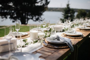 Tahta bir masa, beyaz bir koşucu, beyaz tabaklar ve basit yeşil bir çelenk ile şık bir açık hava düğün masası.