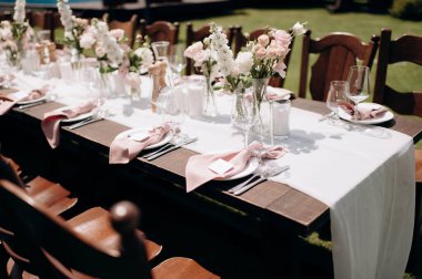 Koyu ahşap bir masa, beyaz bir koşucu, pembe peçeteler ve gür çiçekli süslemeler olan şık bir açık hava düğün masası.