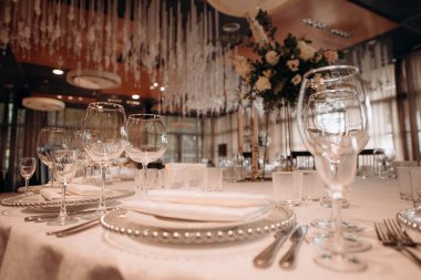 Beyaz masa örtüsü, gümüş boncuklu şarj aletleri, beyaz peçeteler ve uzun çiçek süslemeleri olan şık bir düğün masası.