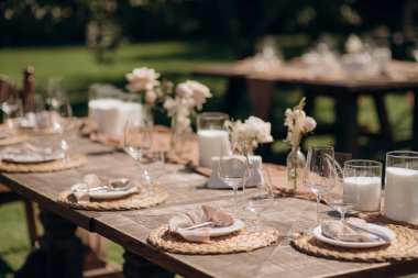 Koyu ahşap bir masa, bej bir koşucu, beyaz tabaklar ve basit çiçek süslemeleri olan kırsal bir açık hava düğün masası.