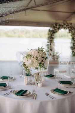 Beyaz masa örtüsü, koyu yeşil peçeteler, beyaz tabaklar ve yemyeşil bir çiçek desenli zarif yuvarlak bir düğün masası.