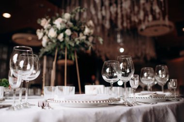 Beyaz masa örtüsü, inci çerçeveli tabaklar ve arka planda bulanık çiçek desenli uzun şarap kadehleri olan zarif bir düğün masası.