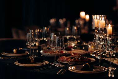 Siyah masa örtüsü, altın çerçeveli tabaklar, koyu peçeteler ve yanan mumlarla dolu gümüş bir şamdan.