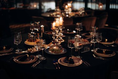 Siyah masa örtüsü, altın çerçeveli tabaklar, koyu peçeteler ve yanan mumlarla dolu gümüş bir şamdan.