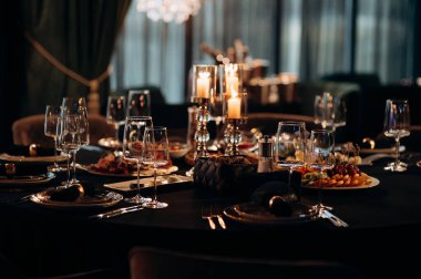 Siyah masa örtüsü, altın çerçeveli tabaklar, koyu peçeteler ve yanan mumlarla dolu gümüş bir şamdan.