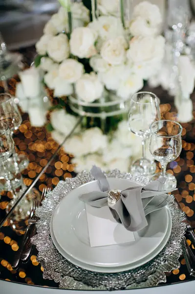 Geniş bir mekanda gümüş tabaklar, gri peçeteler ve kristal avizelerle zarif bir düğün masası.