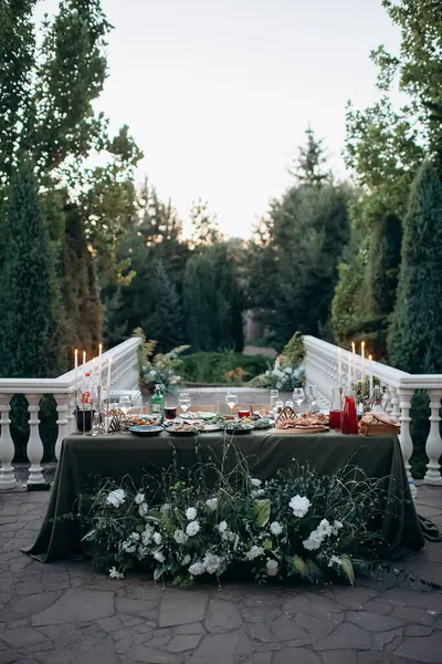 Zeytin yeşili masa örtüsü, yemyeşil çiçek aranjmanı ve mumları olan zarif bir açık hava düğün başkanı.