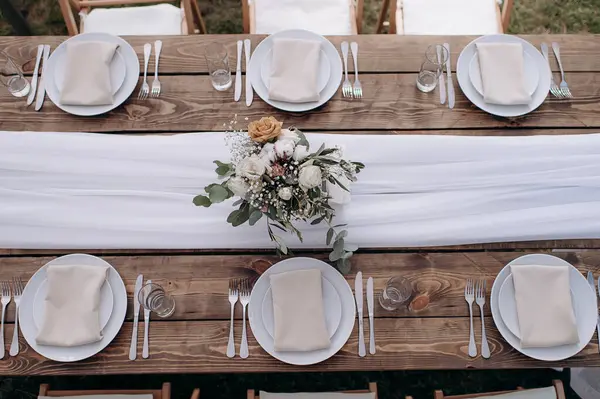Üzerinde ahşap bir masa, beyaz bir koşucu, beyaz plaka ve çiçek desenli bir düğün masası manzarası var.