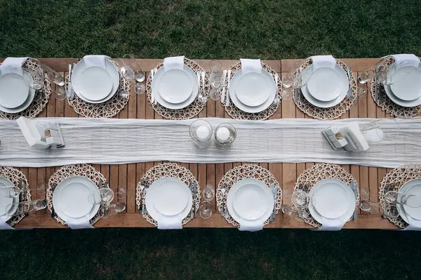 Beyaz tabaklar, peçeteler ve yeşil çimlerin üzerinde beyaz bir koşucuyla zarif bir şekilde kurulmuş ahşap bir düğün masası manzarası.