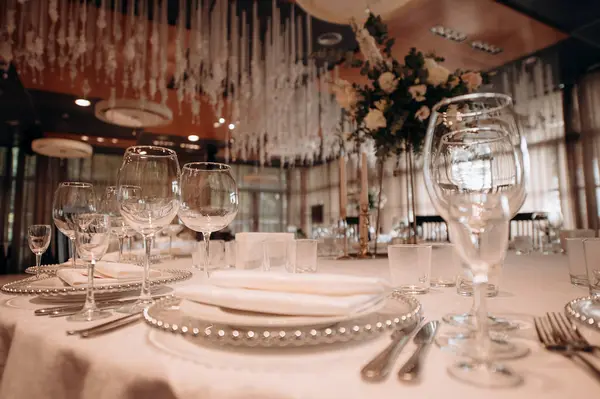 Beyaz masa örtüsü, gümüş boncuklu şarj aletleri, beyaz peçeteler ve uzun çiçek süslemeleri olan şık bir düğün masası.