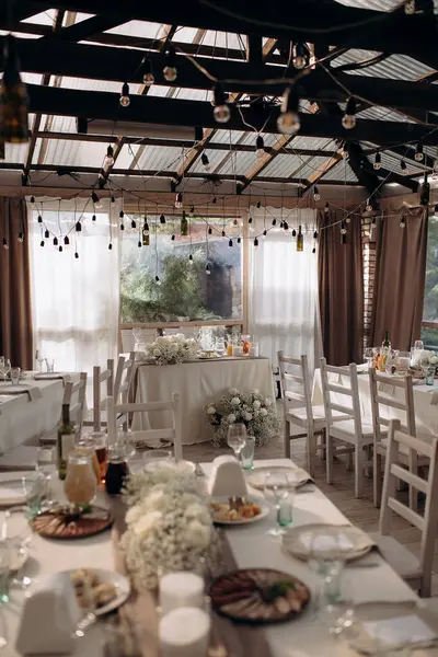 Yuvarlak masalı, beyaz masa örtülü, çiçekli ve sicim lambalı zarif bir düğün resepsiyonu.