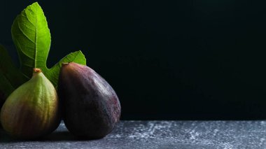 Minimalist kompozisyonda canlı yeşil bir incir yaprağının yanında, toprak ve doğal bir his için dokulu bir yüzeye sahip koyu bir arkaplan üzerine kurulmuş iki olgun incir var..
