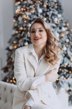 Genç mutlu Avrupalı iş kadını iş yerinde gözlüklü, arkasında süslü bir Noel ağacı olan, kış tatillerinde ev ofisinde çalışan gülümseyen bir kadın. Yumuşak seçici odak.
