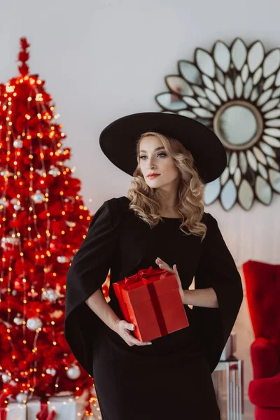 黒のお祝いのドレスと赤いクリスマスツリーの背景に新年の贈り物と帽子の美しいエレガントな女性 柔らかい選択的フォーカス — ストック写真