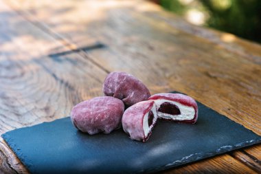 Üç mor mochi topu, siyah bir arka planı olan ahşap bir masada sergilenerek canlı besinler ve doğal elementler arasında güzel bir kontrastlık yaratır. Mochi Asya tatlısı.