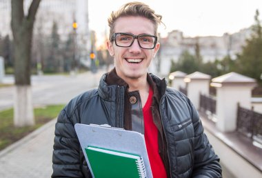 Mutlu bir adam, elinde defterleri olan bir üniversite öğrencisi sokakta yürüyor ve kameraya gülümsüyor.