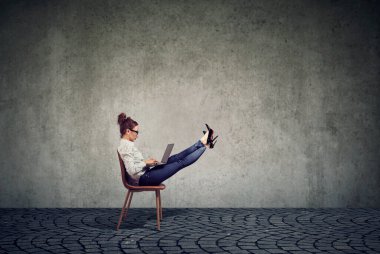 Dizüstü bilgisayarla çalışan güzel bir iş kadını ayakları havada bir sandalyede oturuyor. 