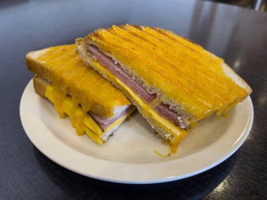 Izgara jambonlu ve peynirli sandviç beyaz tabakta servis edilir ve koyu kahverengi ahşap bir masaya yerleştirilir..