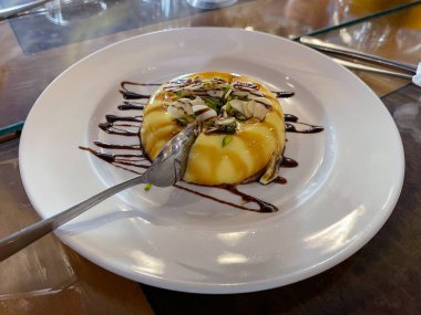 Dilimlenmiş badem ve şam fıstığı ile kaplı beyaz bir tabakta servis edilen karamelli muhallebi..