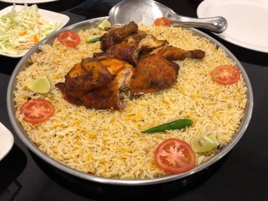 Tavuk biryani büyük çelik bir tabakta servis edilir ve dilimlenmiş domates, yeşil biber ve limon dilimleri ile süslenir..