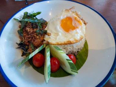 Üzerine kızarmış yumurta ve fesleğen yapraklı kızarmış tavuk konmuş Tayland beyaz pirinci dilimlenmiş salatalık ve kiraz domatesli bir tabakta servis edilir. Tayland yemeği ya da Asya tarzı yemek..