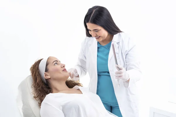 Positive young female patient with curly hair in casual clothes lying on medical equipment and looking at mirror reflection during appointment in aesthetic cosmetology clinic