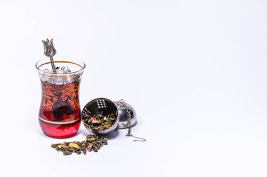 Typical Turkish glass for tea with a teaspoon and tea inside and an open infuser with mango green tea on a white background clipart